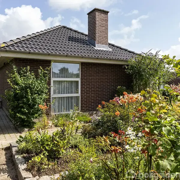 Et rødt murstenshus med mørkt tegltag står omgivet af en blomstrende have. En brolagt sti fører til døren, mens en skorsten hæver sig over taget.