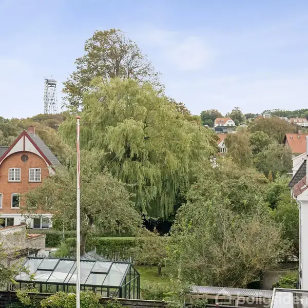 En rød murstensbygning med spidst tag står blandt frodige træer. En flagstang ses i forgrunden, omgivet af en have og et drivhus. I baggrunden er der flere træbeklædte bakker.