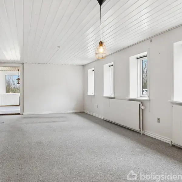 Et tomt rum med lysegrå tæppe og hvide vægge, langs siderne er der tre vinduer og en åbent dør til en balkon. Der hænger en enkelt pendellampe.