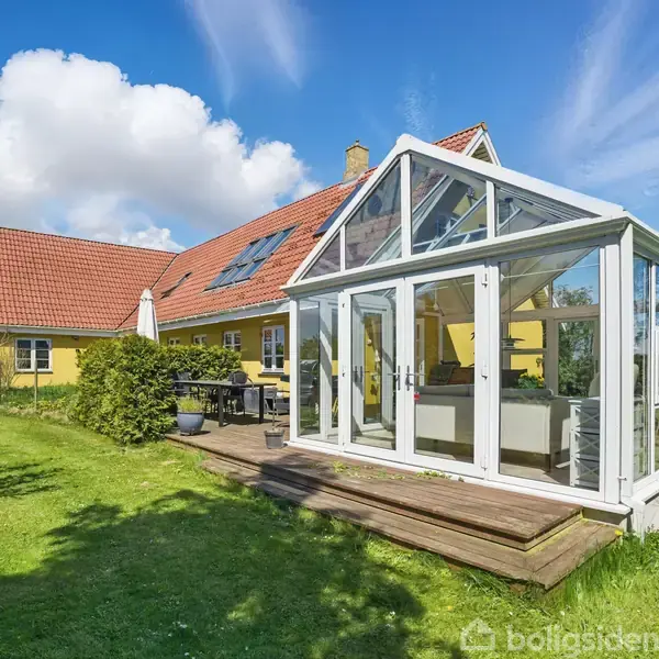 Et gult hus med rødt tegltag har en tilknyttet glaskonstruktion som udestue. Græsplæne og buske omgiver bygningen, mens himlen er blå med få skyer.