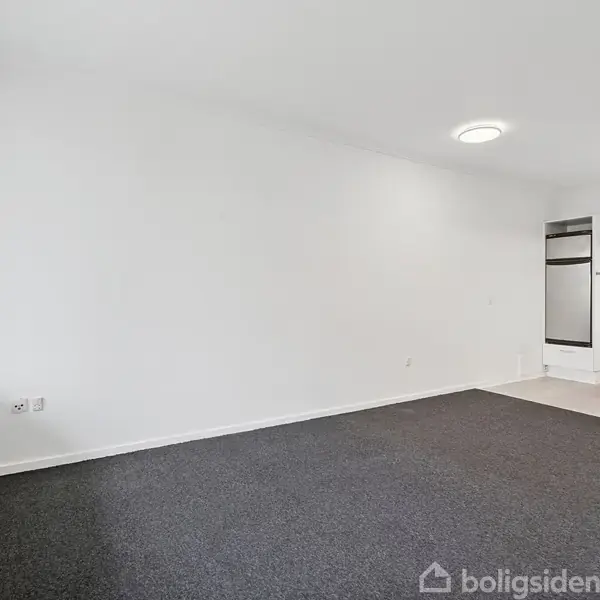 Et tomt rum med gråt tæppe, hvidmalede vægge, og hvidt loft, hvor en del af et køkkenområde med hvide skabe og en ovn ses i baggrunden.