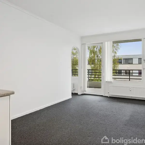 Et lyst værelse med mørkt gulvtæppe har store vinduer, der giver udsigt til en balkon og et grønt træ; køkkenoverflade afgrænser rummet til venstre.