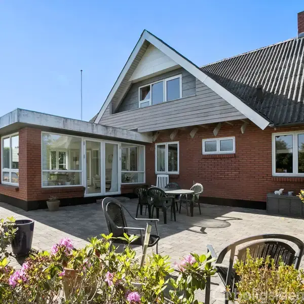 Hus med røde mursten og sort tag, har en terrasse med havemøbler. Sollys skinner på grønne planter og blomster, der omgiver området. Klar blå himmel overalt.