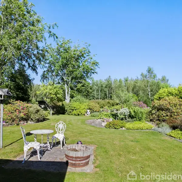 En hvid havebordsæt med stole står på en terrasse i en grøn have omgivet af frodige buske og træer under en klar blå himmel. Fuglehus står ved siden af.