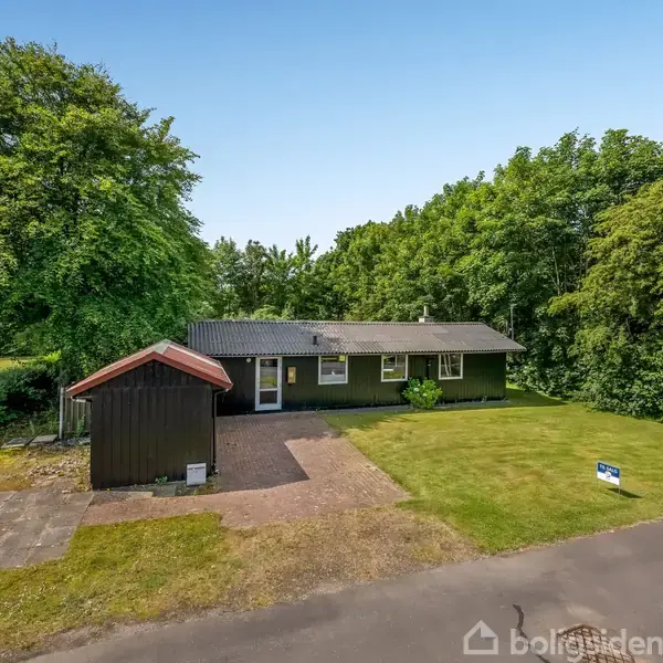 Et sort hus med fladt tag står omgivet af træer. Til venstre ses et skur med rødt tag. Foran huset er en græsplæne og en indkørsel. Skilt i forgrunden.