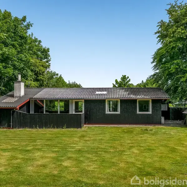 Et sort træhus står stille i en frodig have omgivet af tætte, grønne træer. Græsplænen er velplejet, og der er en lille skur ved siden af huset.