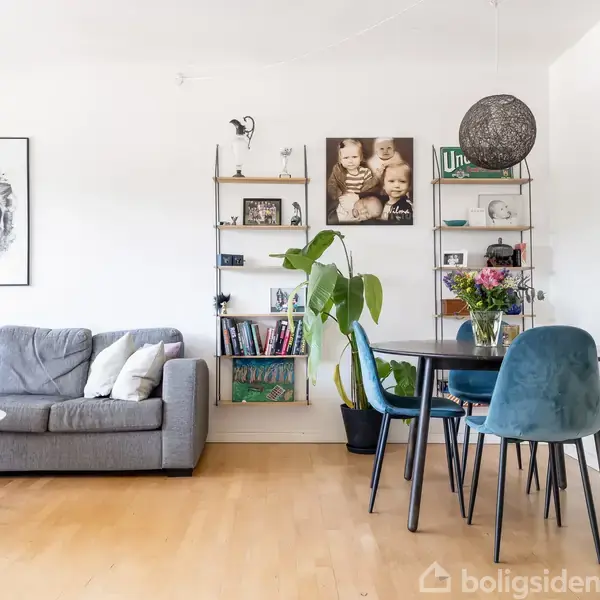 Sofa står til venstre med puder, mens rundt spisebord er omgivet af fire stole til højre. Plante og reol med bøger og dekorationer i baggrunden. Vægkunst og fotos ses.