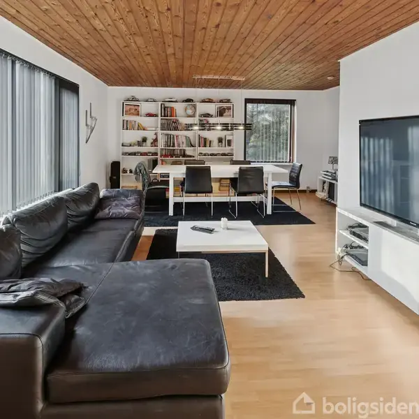 Sort lædersofa står i en stue med trægulv, hvor der er et sofabord og en stor fladskærm. I baggrunden ses et spisebord og reoler med bøger.