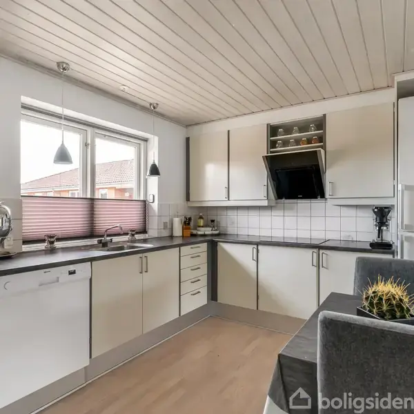 Et moderne køkken med hvide skabe og sorte bordplader. Kaktus på spisebordet. Vindue giver naturligt lys ved opvaskebalje, og hvide fliser dækker væggen over komfuret. Fryser har magneter.