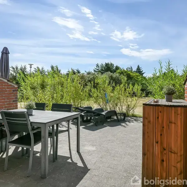 Terrassemøbler står på en flisebelagt terrasse med udsigt til en have. Bord og stole står til venstre, mens omgivelserne består af røde murstensvægge og grøn bevoksning i baggrunden.