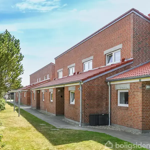 Rækkehuse med murstensfacade står side om side langs en sti. Hver har rødt tegltag og små hvidrammede vinduer. Til venstre er der grøn beplantning under en blå himmel.