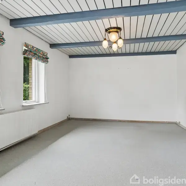 Et tomt værelse med grå tæpper, tre vinduer med farverige gardiner og hvidmalede vægge. En lysekrone hænger fra et loft med blå bjælker.