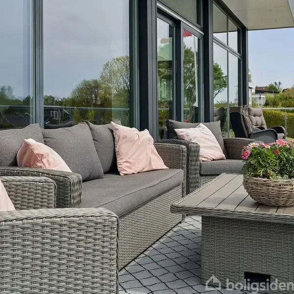 Grå polyrattan sofa og lænestol med lyserøde puder står på en flisebelagt terrasse. Et lavt bord med en blomsterkurv står foran, omgivet af glasvinduer og grønne omgivelser.