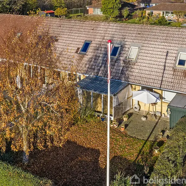 Rækkehus med skråt tag og tagvinduer. Et træ kaster skygge over en lille terrasse med parasol og stol. Haven er omgivet af grønne buske i et boligkvarter.