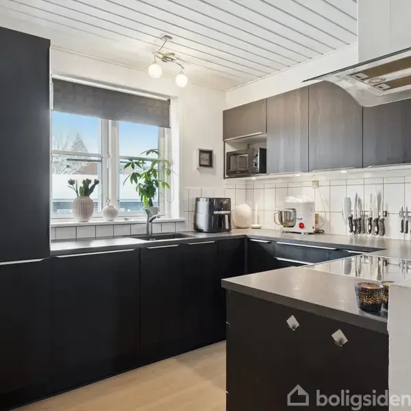 Sort køkken med moderne apparater og planter på vindueskarmen. Knive er arrangeret på væggen under et skab. Lys strømmer ind gennem et vindue, der giver et lyst miljø.