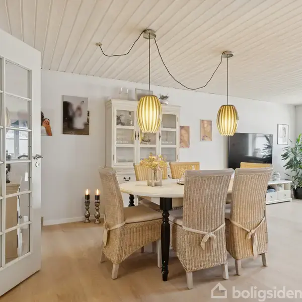 Spisebord med seks stole står i en stue, omgivet af hvide vægge, reol, moderne lamper, og en stor plante nær et vindue. Fjernsyn og sofa ses i baggrunden.