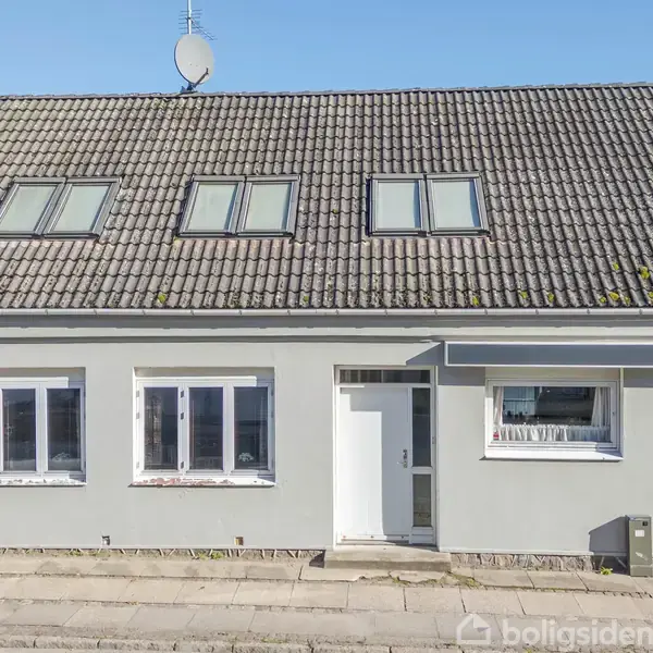 Et gråt murstenshus med skydetag og fem tagvinduer står stille på en brolagt gade. Forneden er der tre store vinduer og en hvid hoveddør.
