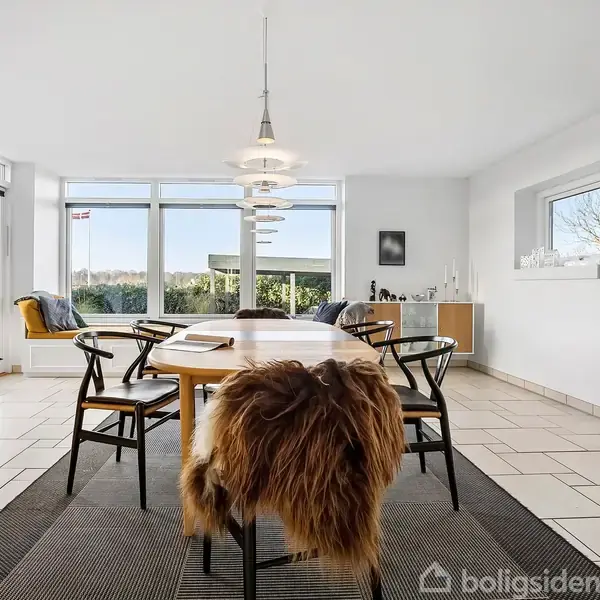 Et spisebord med seks stole er placeret på et tæppe. Der er et stort vinduesparti med udsigt til naturen. Et pelsdækket sæde står forrest. Ingen tekst er synlig.