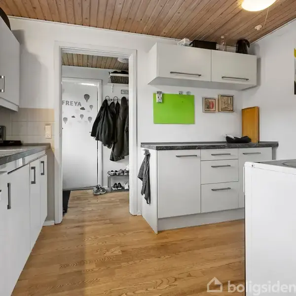 Køkken med hvide skabe og træparketgulv. Skabene har metalliske håndtag og en grøn opslagstavle hænger på væggen. Indgang til en gang med jakker og sko i baggrunden.