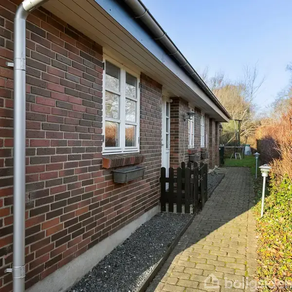 Murstenshus med hvidrammede vinduer og lille sort trækport langs en brolagt sti. Stien er omgivet af buske i sollys, hvilket skaber en hyggelig atmosfære.