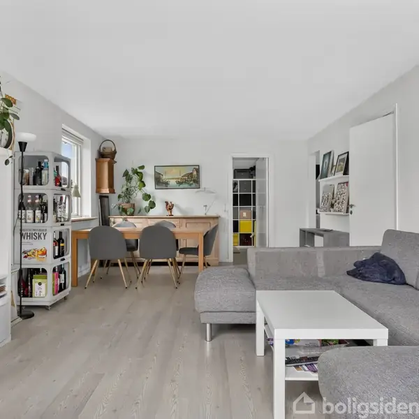 Stue med en grå sofa, sofabord og reol med tv. Et spisebord med fire stole i baggrunden. Grønne planter og billeder på væggene. Lyst trægulv og hvidmalede vægge.