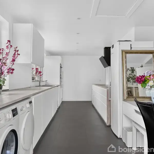 Vaskemaskine og tørretumbler står i et moderne og lyst vaskerum med hvide skabe og sorte gulve. Pyntet med blomster i vindueskarmen og ved et sminkebord med spejl.