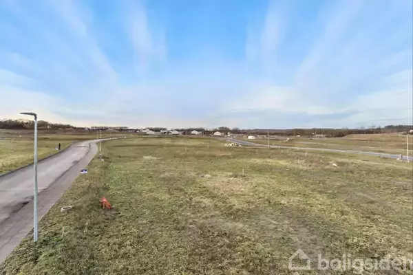 En åben græsmark strækker sig mod en horisont med spredte bygninger i det fjerne. En brolagt sti løber langs venstre side med enkelte gadelygter. Himlen er klar.