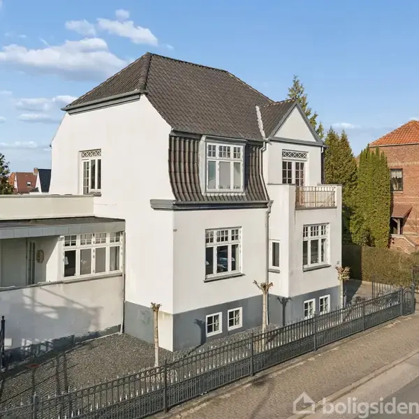Et hvidt, toetagers hus står ved en vej omgivet af et stakit og en have. Der er en balkon, og taget er mørkegråt. I baggrunden ses flere huse.