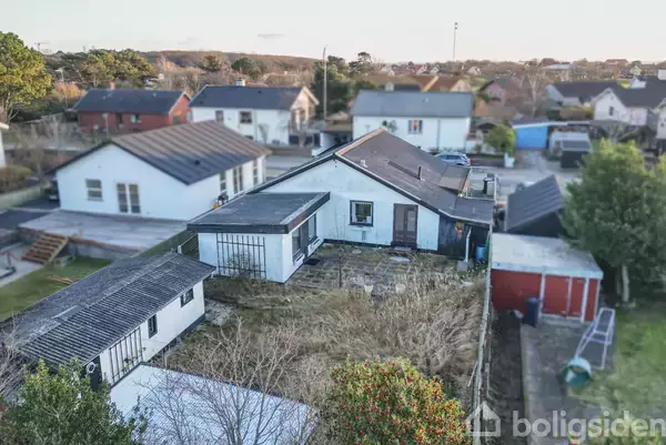 Et hvidt hus med fladt tag står i en have omgivet af buske og græs. Andre huse ses i baggrunden i et beboelseskvarter.