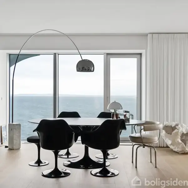 Et moderne spisebord sæt med sorte stole står foran store vinduer med havudsigt. En stilfuld lampe hænger over bordet, og rummet har lyse trægulve med minimalistisk indretning.