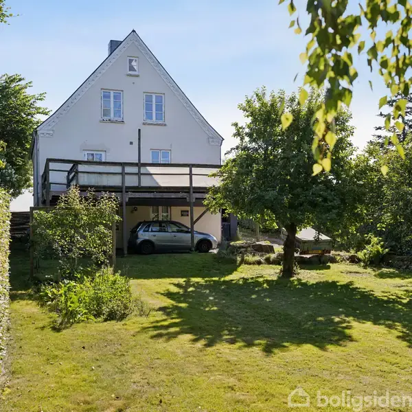 Et hvidt, etplanshus står i en grøn have. En bil er parkeret under en overdækket terrasse. Træer og buske omgiver huset, hvilket skaber en rolig atmosfære.