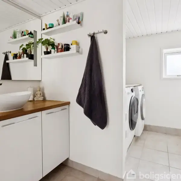 Håndvasken står på et træbord med skuffer, omgivet af hylder med planter og toiletsager. En vaskemaskine og tørretumbler befinder sig i det lyse, flisebelagte rum.