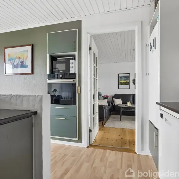 Køkken med hvide og grønne skabe, hvor mikroovn og genstande er placeret. Gennem åben dør ses stue med sofa og sofabord. Trægulv og hvidt loft dominerer rummet.