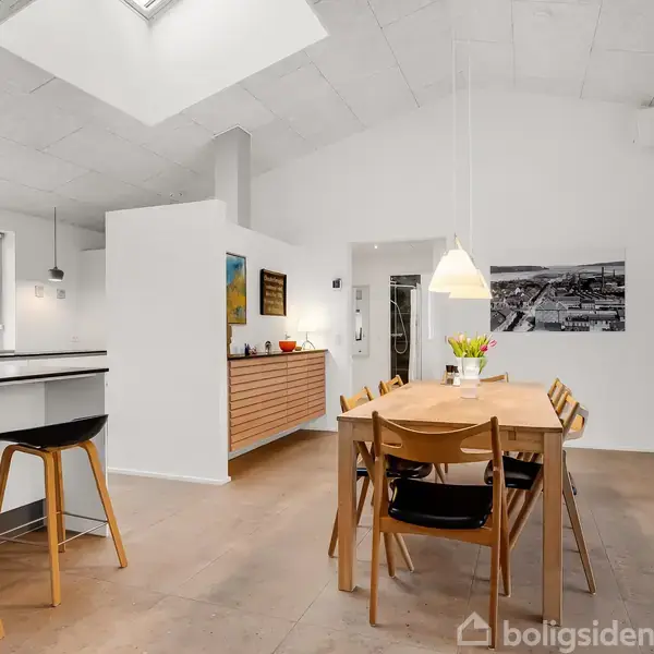 Et spisebord med seks stole står i et lyst rum. To barstole ved en køkkenbar ses til venstre. Væggene er hvide, og der hænger kunst og lamper.