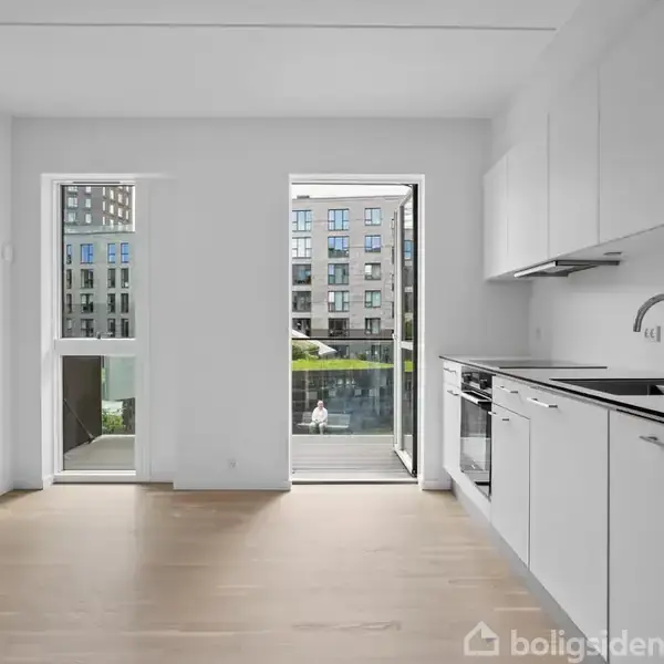Et moderne køkken med hvide skabe og apparater, åbent mod en lys stue. Et stort vindue og en dør fører ud til en balkon. Udsigt til lejlighedskompleks.