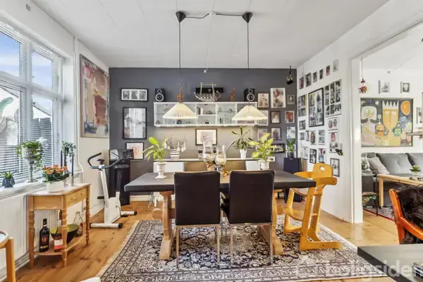 En spisestue med et sort spisebord omgivet af sorte og gule stole står på et orientalsk tæppe. Væggene er beklædt med kunstplakater og hylder. Vinduer til venstre.