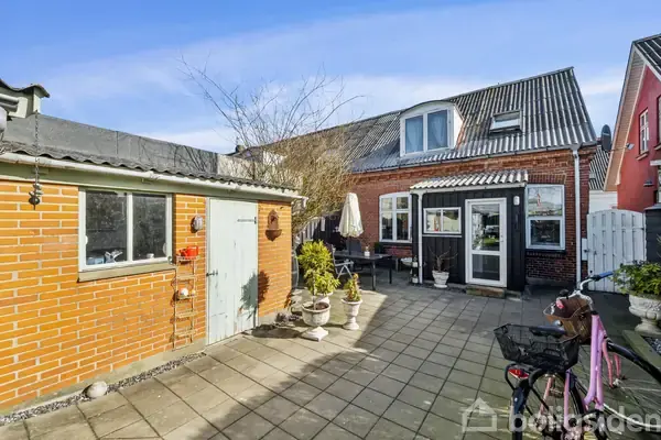 En cykel står parkeret på en flisebelagt gårdsplads. I baggrunden ses et rødt murstenshus med et stort vindue og en dør. Alt sammen under en blå himmel.