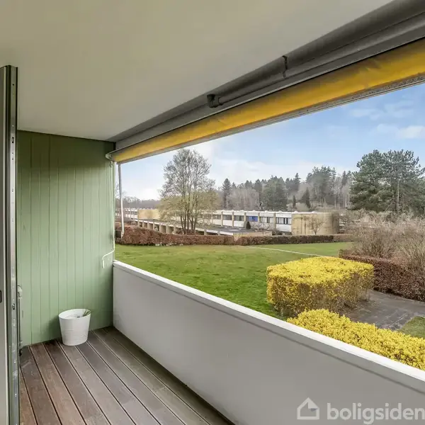 En overdækket altan med åbent glasdør vises med en grøn væg, træplanker, og en hvid potte. Udsigt til have med buske, træer og græsarealer.