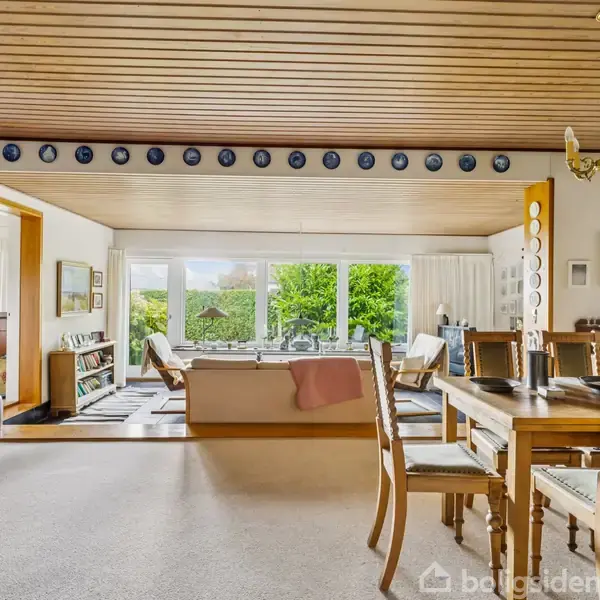 Et bord med fire stole står på en flisebelagt terrasse ved siden af et murstenshus. Grønne buske og træer omgiver området, mens himlen er blå og klar.