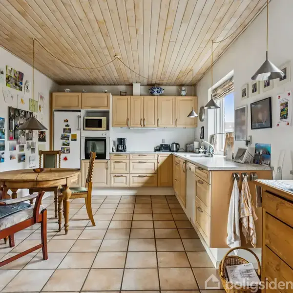 Køkken med træloft, lyse træskabe og beige fliser. Rundt bord med stole står op ad væg med billeder og tegninger. Vinduer og hængende lamper giver lys.