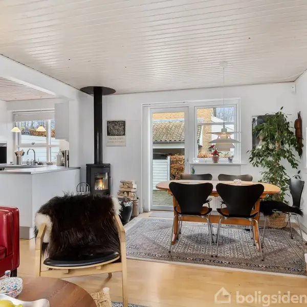 Stue med spisebord og fire stole omkring det. Brændeovn ved siden af et vindue, der slipper dagslys ind. Omgivet af lyse farver og moderne indretning.