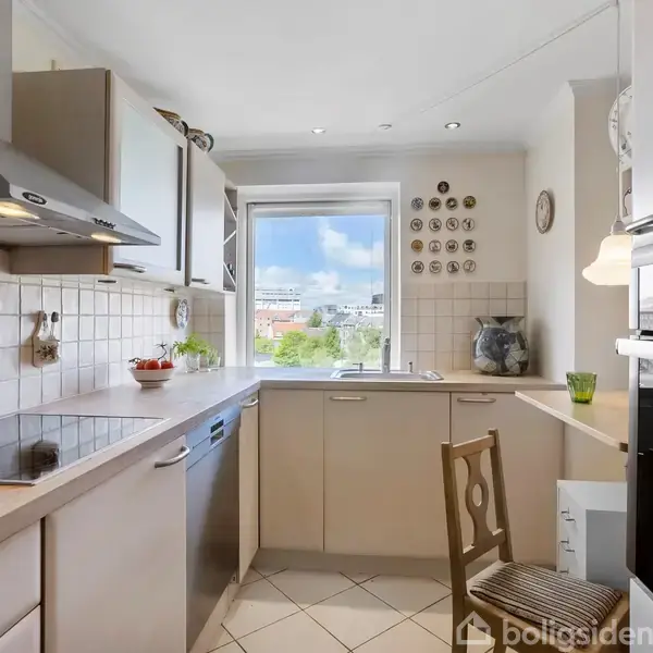 Køkken med moderne apparater. En ovn, opvaskemaskine og kogeplader optager pladsen, mens en træstol står ved et vindue. Flisevæg og hvidt skab kompletterer det lyse, enkle design.