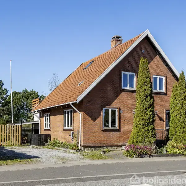 Et rødt murstenshus med skråt tag står ved en vej. Huset har to høje, smalle træer foran og en indkørsel til venstre. Det er en klar dag.