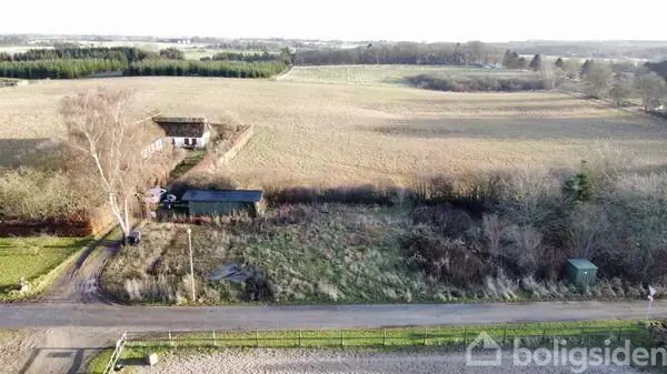 En bygning står stille omgivet af en stor, åben mark under en klar himmel. I forgrunden løber en vej, og vegetationen er sparsom.