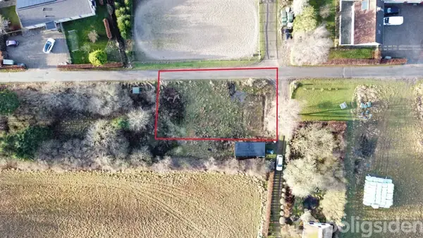 Et rektangulært jordareal, markeret med rød linje, set fra oven, omgivet af bebyggelse og marker med beplantede træer og buske i et tætliggende boligområde.