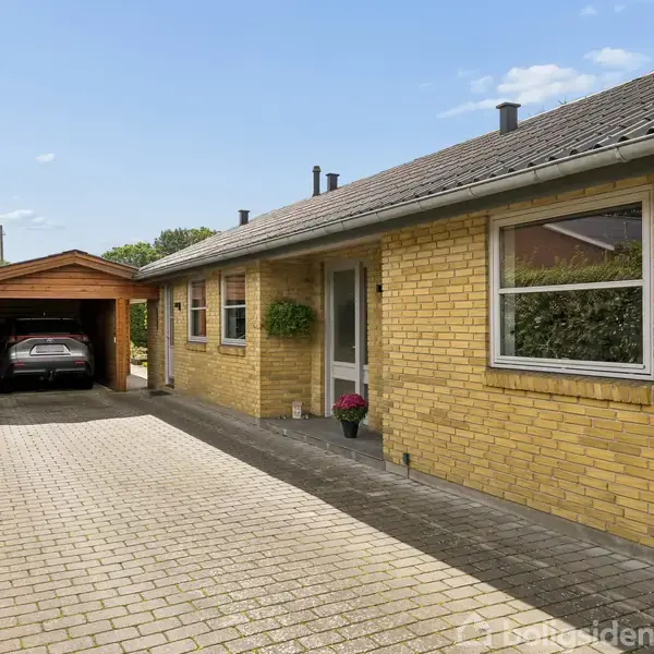 En gul murstenshus står stille langs en brolagt indkørsel. En bil er parkeret i en carport ved siden af huset, omkranset af grønne hække under en blå himmel.