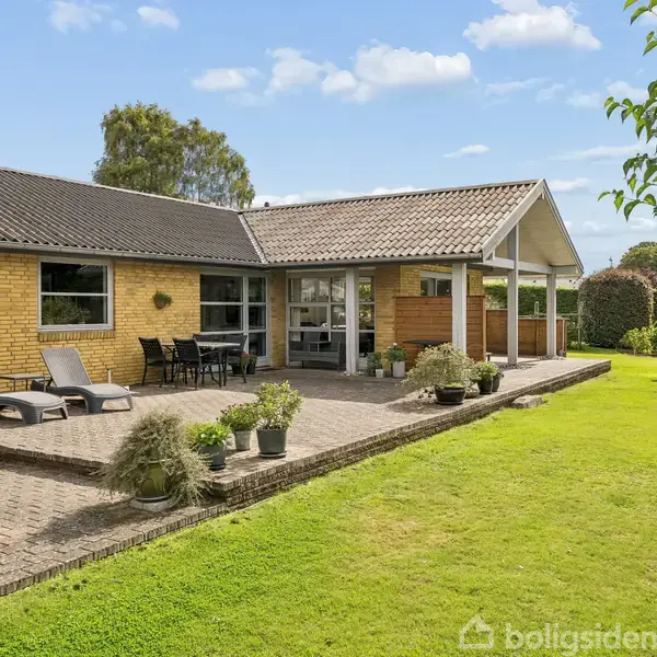 Hus i mursten med overdækket terrasse, hvor liggestole og bord er placeret, omgivet af velplejet græsplæne og træer i en solrig have.