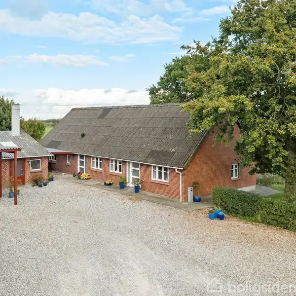 Rødt murstenshus med skråt tag står stille omgivet af grusareal. En carport huser to biler til venstre, og store træer samt græs udgør baggrunden.