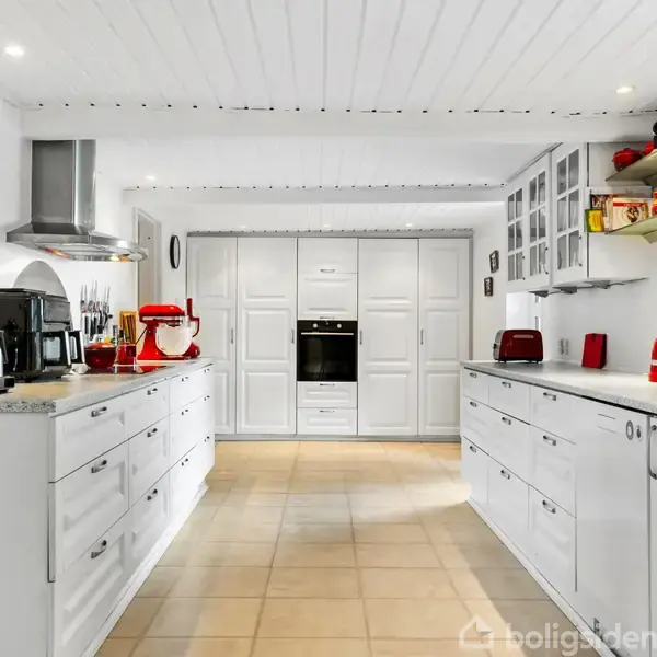 Et moderne køkken med hvide skap. Bordplader har køkkenmaskiner som kaffemaskine og brødrister. Rummet har flisegulv, indbyggede ovne og åben hylde med dekorationer.