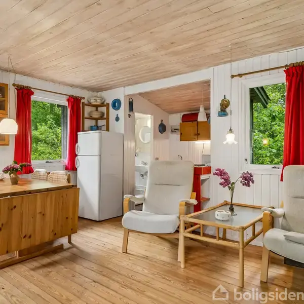 Stue med træmøbler og to lænestole ved et bord med blomster. Et spisebord står ved vinduer med røde gardiner. I baggrunden er et køkken og et hvidt køleskab.